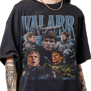May include: Dark gray t-shirt featuring a collage of images of a person, with the text "VALARR Targaryen" in blue and white. The design includes lightning bolts and a vintage aesthetic.