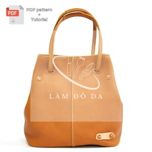 May include: A two-tone leather tote bag with a light brown upper and a darker brown lower section. The bag has two handles and decorative stitching. The text "LÀM ĐỒ DA" is visible on the front. A PDF pattern and tutorial icon are in the upper left.