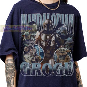 May include: Navy blue t-shirt featuring a graphic design with the text "MANDALORIAN" and "GROGU". The design includes images of characters from the Star Wars series, including The Mandalorian and Grogu. The shirt has a crew neck and short sleeves.