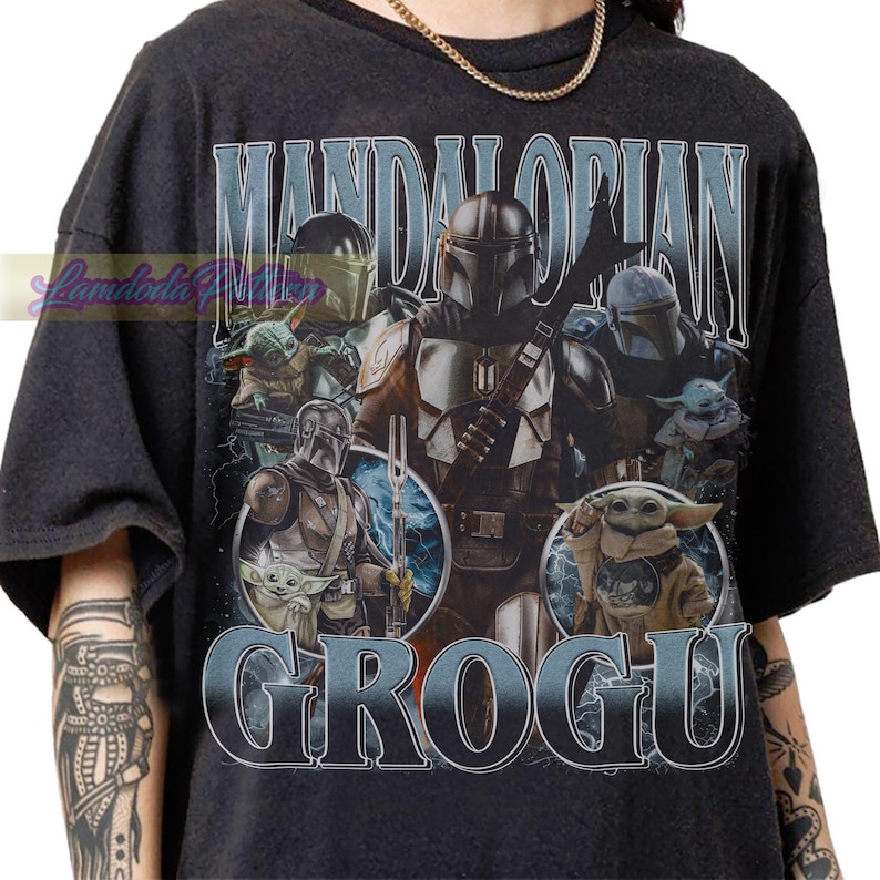 May include: Dark gray t-shirt featuring a graphic of characters from The Mandalorian series, including the Mandalorian and Grogu. The design includes the words "MANDALORIAN" and "GROGU" in a distressed, vintage style.