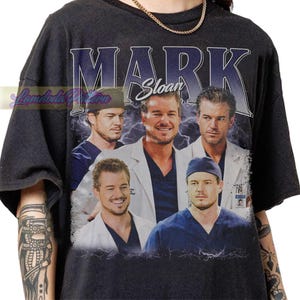 May include: Black t-shirt featuring a collage of images of a man in various poses, with the name "MARK Sloan" in large, stylized text above the images. The design has a vintage, distressed look with lightning bolt accents.