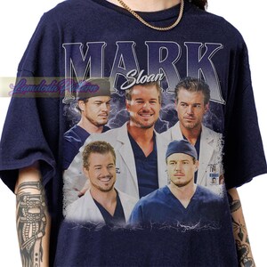 May include: Navy blue t-shirt featuring a collage of images of a man, with the name "MARK Sloan" in large white letters. The images are set against a background of lightning bolts.