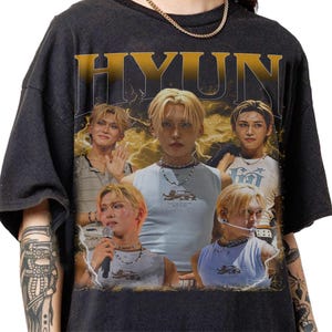 May include: Black t-shirt featuring a graphic design with the name "HYUN" in gold lettering. The design includes multiple images of a person with blonde hair, wearing various outfits. The background has a vintage aesthetic with lightning bolts.