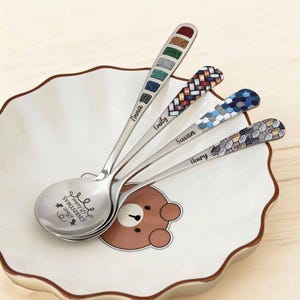 Puede incluir: Cuatro cucharas de acero inoxidable con mangos decorativos sobre un plato blanco con diseño de oso. Cada cuchara tiene un diseño de mango diferente, incluyendo purpurina de colores, patrones tejidos y formas geométricas. Los nombres están grabados en los mangos.
