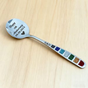 May include: A stainless steel spoon with the text "Happy 11th Anniversary 08.09.2014" and a heart symbol engraved on the spoon's bowl. The handle features a series of colorful glitter-filled squares. The initials "G&D" are also engraved.