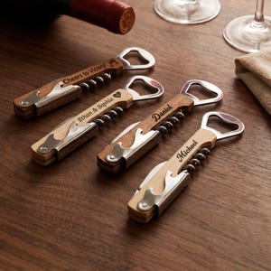 Op de afbeelding: Vier gepersonaliseerde houten wijnopeners met zilveren accenten en flesopeners. Elke opener heeft een andere naam of zin gegraveerd op de houten handgreep: "Cheers to Victory!", "Ethan & Sophia", "Daniel" en "Michael".