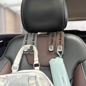 May include: Car seat headrest hooks with the text "No Mess", "No Stress", "Travel Light", and "Live Bright". A white purse and a light blue water bottle hang from the hooks. The hooks are gray and brown.