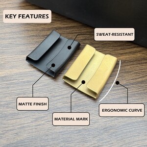 May include: Two rectangular stainless steel keepers with a matte finish, one black and one gold. Each piece features an ergonomic curve and is labeled with the text STAINLESS STEEL. Text labels indicate key features: KEY FEATURES, SWEAT-RESISTANT, MATTE FINISH, MATERIAL MARK, and ERGONOMIC CURVE.