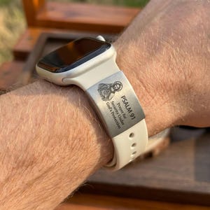 May include: A white silicone smartwatch band featuring a rectangular silver-toned metal charm engraved with an illustration of Jesus and the text PSALM 91 Prayer for Security Under God's Protection. The band has a smooth finish and buckle holes.