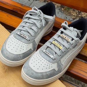 Könnte beinhalten: Ein Paar grau-weiße Sneaker mit silbernen Akzenten und schwarzem Besatz. Die Sneaker haben graue Schnürsenkel und kleine Metallanhänger mit Text. Ein Anhänger lautet "KEEP MOVING" und ein anderer "I CAN DO IT".