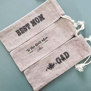 Puede incluir: Tres bolsas de lino beige con cordón y texto negro. La bolsa superior dice "BEST MOM", la bolsa del medio dice "To the Best Mom Alice", y la bolsa inferior muestra "G&D" con un diseño floral.
