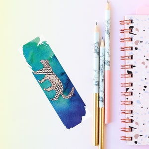 May include: A watercolour bookmark with a blue and green background featuring a black and white patterned animal. Three pencils with marble and pink designs are next to a pink and white speckled notebook.