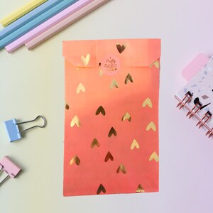 Peut inclure: Une enveloppe orange et rose avec des découpes de cœurs dorés et un petit autocollant. L'enveloppe est entourée de crayons colorés, de pinces à reliure et d'un carnet à spirales avec un motif terrazzo.