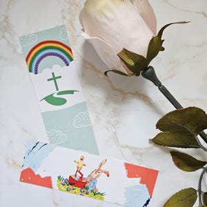 May include: Two illustrated bookmarks and a pink rose. One bookmark has a rainbow, cross, and green landscape. The other depicts a rabbit and chick with an Easter theme. The background is a marble pattern. The rose is a pale pink.