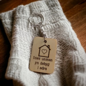 May include: A wooden keychain with a house design and the text "Does unman yn debyg i adra". The keychain is attached to a silver key ring.