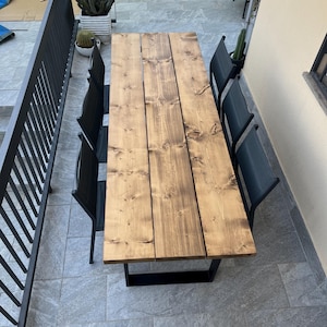Pu&ograve; includere: Un tavolo da pranzo rettangolare in legno per esterni con una macchia scura, sostenuto da una base in metallo nero. Sei sedie nere circondano il tavolo. Il tavolo misura circa 183 cm di lunghezza.
