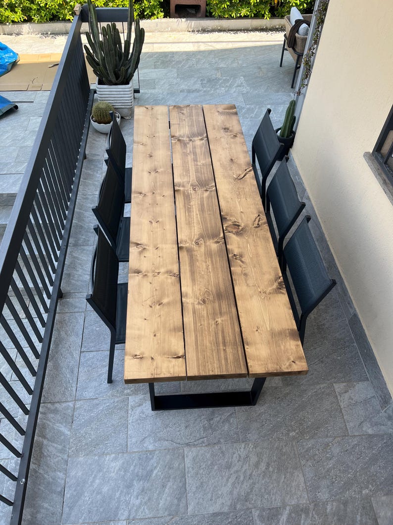 Pu&ograve; includere: Un tavolo da pranzo rettangolare in legno per esterni con una macchia scura, sostenuto da una base in metallo nero. Sei sedie nere circondano il tavolo. Il tavolo misura circa 183 cm di lunghezza.