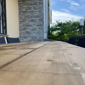 Pu&ograve; includere: Un lungo tavolo in legno marrone chiaro con venature visibili. Sedie nere sono posizionate lungo il lato del tavolo. Lo sfondo include un muro di pietra, una finestra con sbarre nere e una vegetazione lussureggiante sotto un cielo blu.