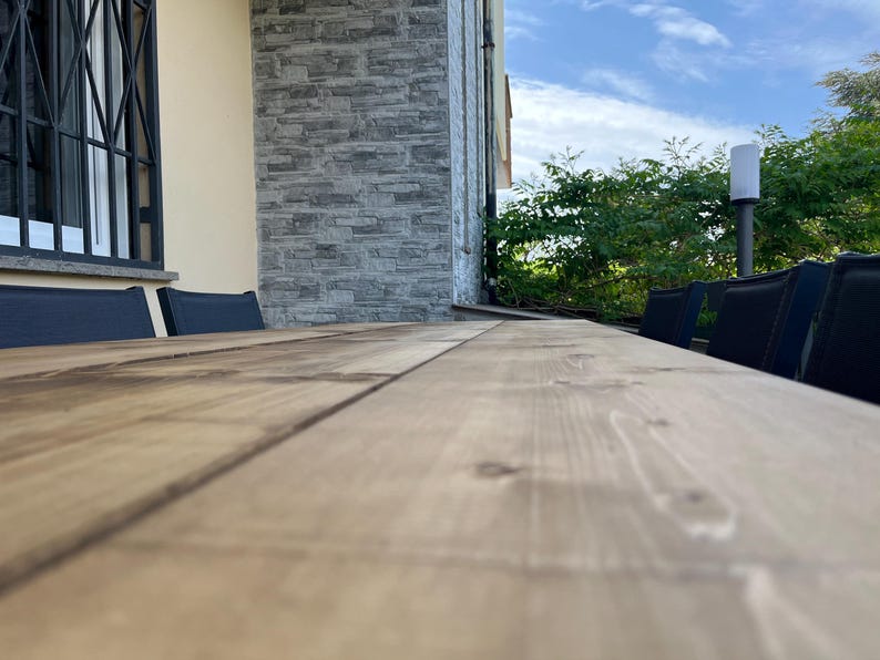 Pu&ograve; includere: Un lungo tavolo in legno marrone chiaro con venature visibili. Sedie nere sono posizionate lungo il lato del tavolo. Lo sfondo include un muro di pietra, una finestra con sbarre nere e una vegetazione lussureggiante sotto un cielo blu.