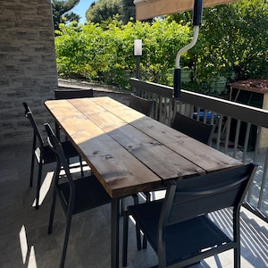 Pu&ograve; includere: Un tavolo da pranzo esterno in legno scuro, abbinato a sei sedie in metallo nero. Il tavolo rettangolare, probabilmente in legno riciclato, &egrave; circondato da sedie dal design moderno. La scena &egrave; ambientata su un patio con una tenda da sole retrattile.
