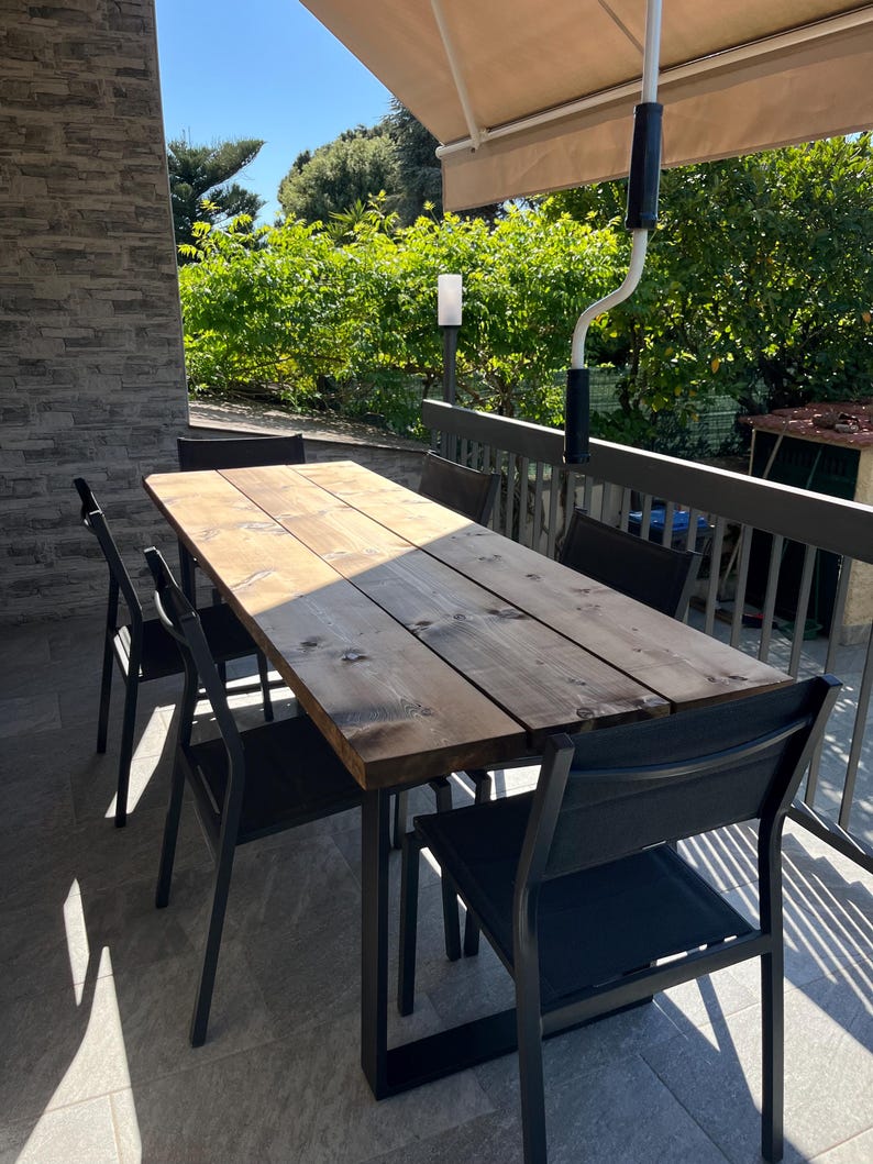Pu&ograve; includere: Un tavolo da pranzo esterno in legno scuro, abbinato a sei sedie in metallo nero. Il tavolo rettangolare, probabilmente in legno riciclato, &egrave; circondato da sedie dal design moderno. La scena &egrave; ambientata su un patio con una tenda da sole retrattile.