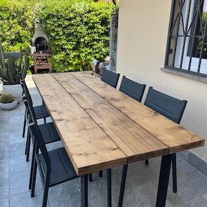 Pu&ograve; includere: Un tavolo da pranzo rettangolare in legno per esterni con finitura naturale, abbinato a sei sedie in metallo nero. Il tavolo ha una tinta marrone scuro e poggia su gambe in metallo nero.