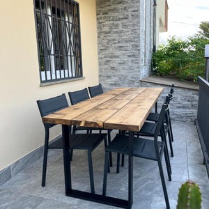Pu&ograve; includere: Set da pranzo all'aperto con un tavolo rettangolare in legno (circa 200 cm di lunghezza) e sei sedie nere. Il tavolo ha gambe in metallo nero. Il set si trova su un patio piastrellato, con un edificio e una finestra sullo sfondo.