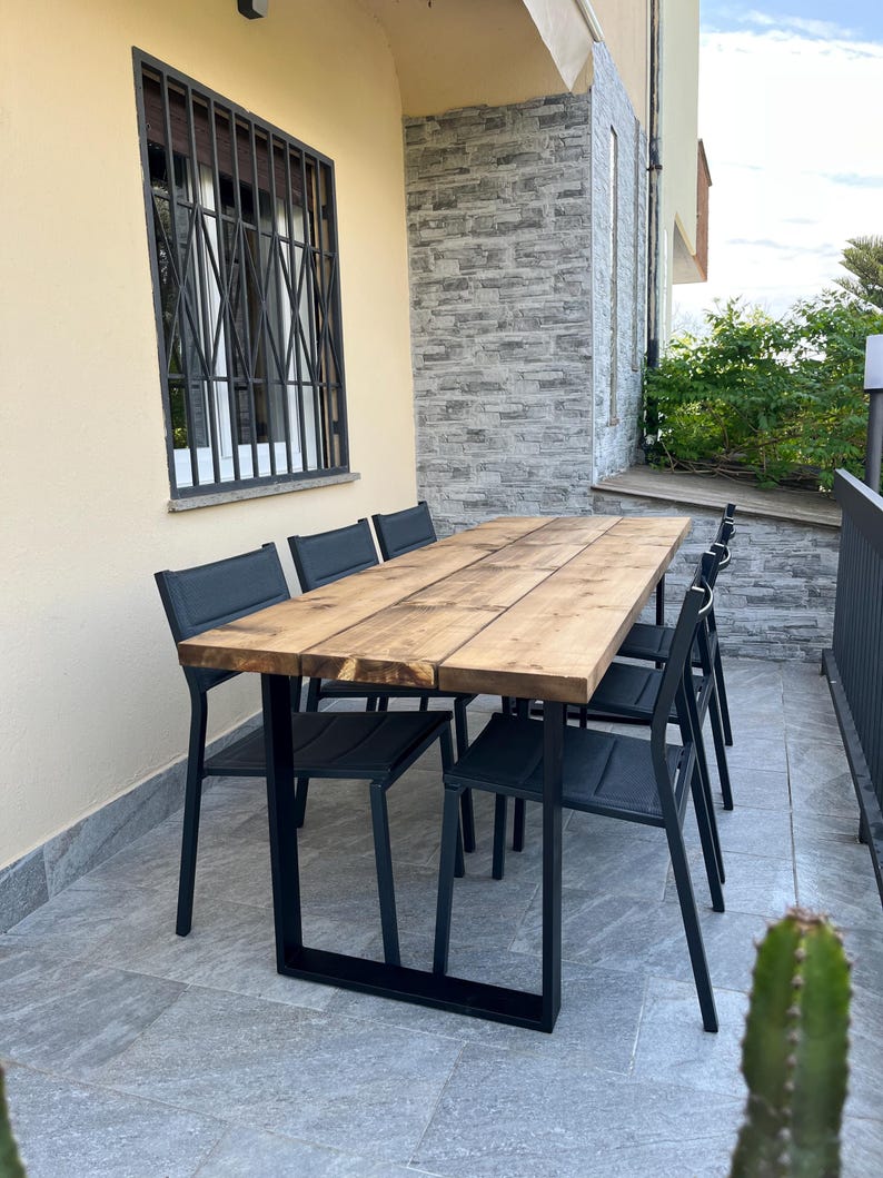 Pu&ograve; includere: Set da pranzo all'aperto con un tavolo rettangolare in legno (circa 200 cm di lunghezza) e sei sedie nere. Il tavolo ha gambe in metallo nero. Il set si trova su un patio piastrellato, con un edificio e una finestra sullo sfondo.