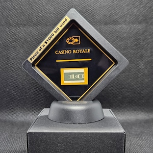 May include: A black display case with a gold-trimmed, diamond-shaped frame. The frame holds a piece of a $1000 bill and the text "CASINO ROYALE". The case sits on a black base and a black box.