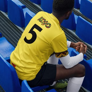 Pode incluir: Camisola de futebol amarela com o número 5 em preto nas costas. A camisola tem o nome "Kojo" em letras coloridas e um design estampado nas mangas. A pessoa está sentada em assentos azuis do estádio.