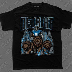 May include: Black t-shirt with a graphic print of a football player in a blue uniform with the word "DETROIT" in large white letters above the player. The player is standing in front of a cityscape with three lions in front of him.