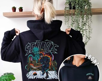 May include: Black hoodie with a graphic print on the back of a hockey player with a kraken on the ice. The front of the hoodie has a small embroidered graphic of a skull and crossbones with the word "Seattle" above it.