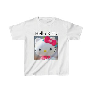 以下が含まれることがあります： 「ハローキティ」の文字とハローキティのキャラクターの画像がプリントされた白いTシャツ。キャラクターは白い頭、ピンクのリボン、ピンクのドレスを着ています。Tシャツは柔らかい素材で作られています。