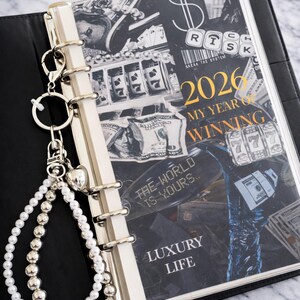 May include: A black planner with a silver binder and a decorative charm. The cover features a collage of money and the text "2026 My Year of Winning" and "Luxury Life". A charm with pearls and silver beads hangs from the binder.