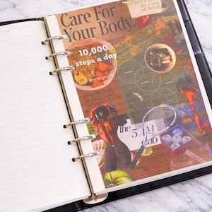May include: Open black leather-bound planner with lined pages and a collage insert. The insert features text that reads "Care For Your Body" and "10,000 steps a day." The collage includes images of food, tennis balls, and a tennis racket.