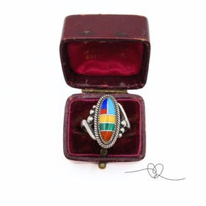 K&ouml;nnte beinhalten: Ein silberner Ring mit einem ovalen Stein, der in der Mitte eingesetzt ist. Der Stein besteht aus mehreren bunten Steinen, darunter Rot, Orange, Gelb, Gr&uuml;n, Blau und T&uuml;rkis. Der Ring befindet sich in einer roten Samt-Ringbox.