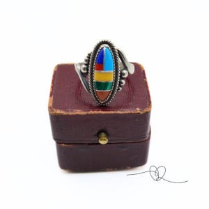 K&ouml;nnte beinhalten: Ein silberner Ring mit einem ovalen Stein, der in der Mitte eingesetzt ist. Der Stein besteht aus mehreren Farben von T&uuml;rkis, Rot, Gelb, Gr&uuml;n und Blau. Der Ring befindet sich in einer kleinen roten Schachtel.