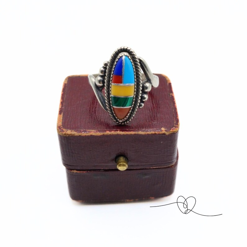 K&ouml;nnte beinhalten: Ein silberner Ring mit einem ovalen Stein, der in der Mitte eingesetzt ist. Der Stein besteht aus mehreren Farben von T&uuml;rkis, Rot, Gelb, Gr&uuml;n und Blau. Der Ring befindet sich in einer kleinen roten Schachtel.