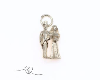 May include: Silver charm featuring a bride and groom figurine. The bride is wearing a long gown and the groom is wearing a top hat and suit. The charm is perfect for adding a touch of romance to any jewelry piece.