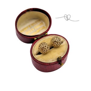 May include: Two gold spherical earrings with an intricate openwork filigree pattern resting inside an oval-shaped, deep red velvet-lined jewelry box. The interior lid of the box features gold lettering, and a small heart line drawing is visible.