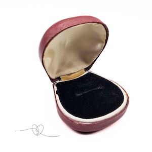 May include: An open, burgundy-colored ring box with a black velvet interior and a cream-colored satin lining. The box is heart-shaped and has a hinged lid. A simple line drawing of a heart is in the lower left corner.