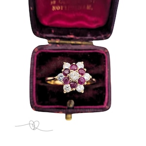 Op de afbeelding: Een gouden ring met een bloemmotief, met een centrale diamant omringd door rode robijnen en kleinere diamanten. De ring wordt gepresenteerd in een vintage bordeauxrode fluwelen sieradendoos. Het ontwerp van de ring doet denken aan een bloem.