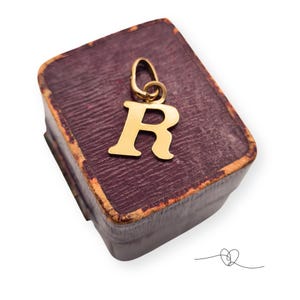 Könnte beinhalten: Ein goldfarbener Anhänger mit dem Buchstaben "R" und einer Schlaufe für eine Kette, der auf einer alten, weinroten Schmuckschatulle liegt. Die Schatulle hat eine abgenutzte, strukturierte Oberfläche und einen goldfarbenen Rand. Der Anhänger ist der Blickfang.