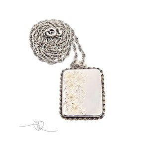 May include: A silver-toned rectangular pendant necklace with a floral design. The pendant has a textured border and a chain. The floral design is on the right side of the pendant. The necklace is on a white background.
