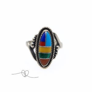 K&ouml;nnte beinhalten: Ein silberner Ring mit einem ovalen Stein, der in der Mitte eingesetzt ist. Der Stein besteht aus mehreren Farben von Einlegearbeiten, darunter Rot, Orange, Gelb, Gr&uuml;n, Blau und T&uuml;rkis.