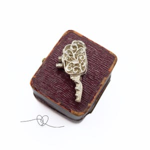 May include: A silver key charm with an intricate design, displayed in a burgundy velvet box.