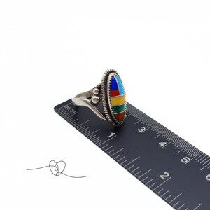 K&ouml;nnte beinhalten: Ein silberner Ring mit einem mehrfarbigen ovalen Stein, der in der Mitte eingesetzt ist. Der Stein besteht aus T&uuml;rkis, Rot, Gelb, Gr&uuml;n und Blau. Der Ring wird an einem Lineal gemessen und zeigt, dass er etwas mehr als 2,5 cm im Durchmesser hat.