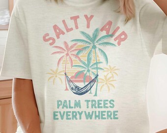 May include: Cream-colored t-shirt with the text "SALTY AIR PALM TREES EVERYWHERE" in pink, blue, and yellow. The graphic features palm trees and a hammock. The shirt is a casual, relaxed fit.
