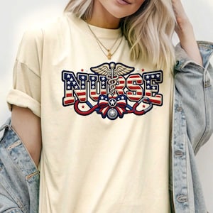 Può includere: T-shirt color crema con un design patriottico. La parola "NURSE" è visualizzata in rosso, bianco e blu, con stelle e strisce. Un simbolo di caduceo, uno stetoscopio e un fiocco completano il design.