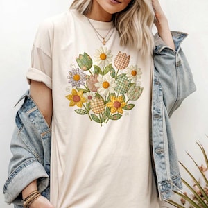 May include: Cream-coloured t-shirt with a floral bouquet design. The bouquet features daisies, tulips, and other flowers in various colours and patterns, including gingham and floral prints. The shirt has short sleeves and a relaxed fit.